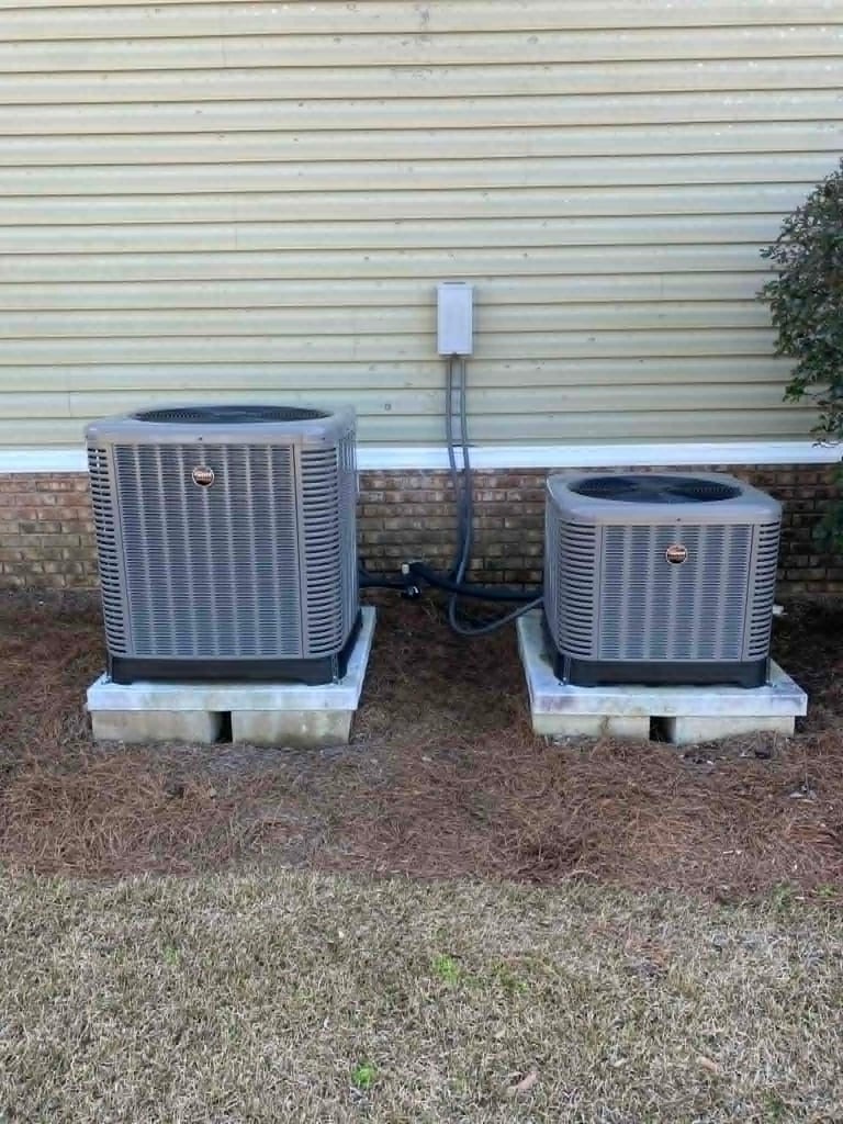 Two air conditioning units mounted on concrete pads outside a brick and vinyl sided house with electrical disconnect box mounted between them