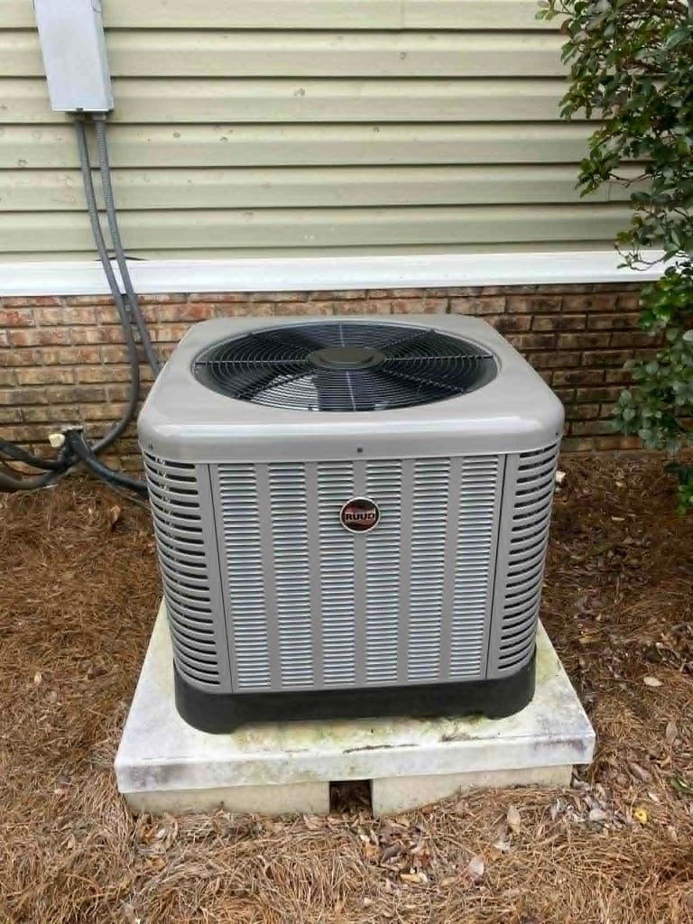 Outdoor air conditioning unit mounted on concrete pad against brick foundation with green siding and climbing ivy above