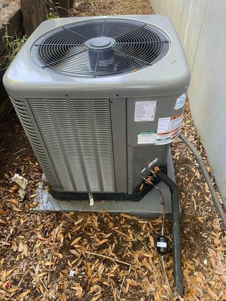 Outdoor HVAC unit installed on mulch ground with conduit tubing and white siding visible in background
