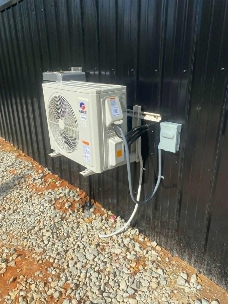 Heat pump unit with compressor and fan mounted on dark corrugated metal wall above gravel ground
