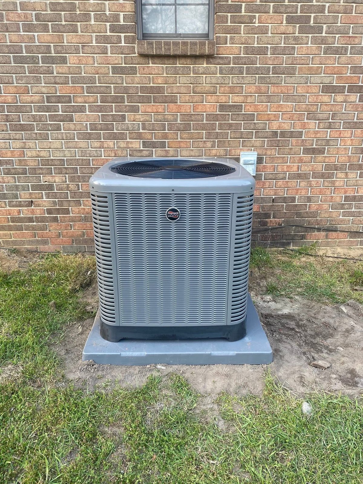 Air conditioning unit installed on concrete pad against brick wall with lawn and basement window visible